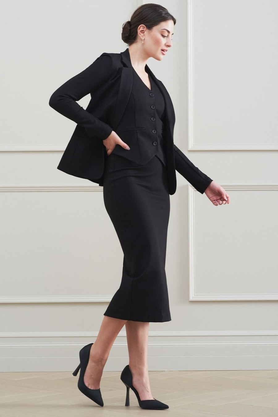 Woman wearing a black suit standing against a light-colored wall.