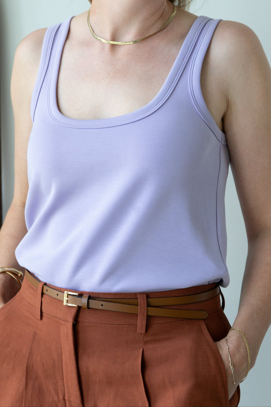 Woman wearing a light purple tank top and brown pants against a plain background