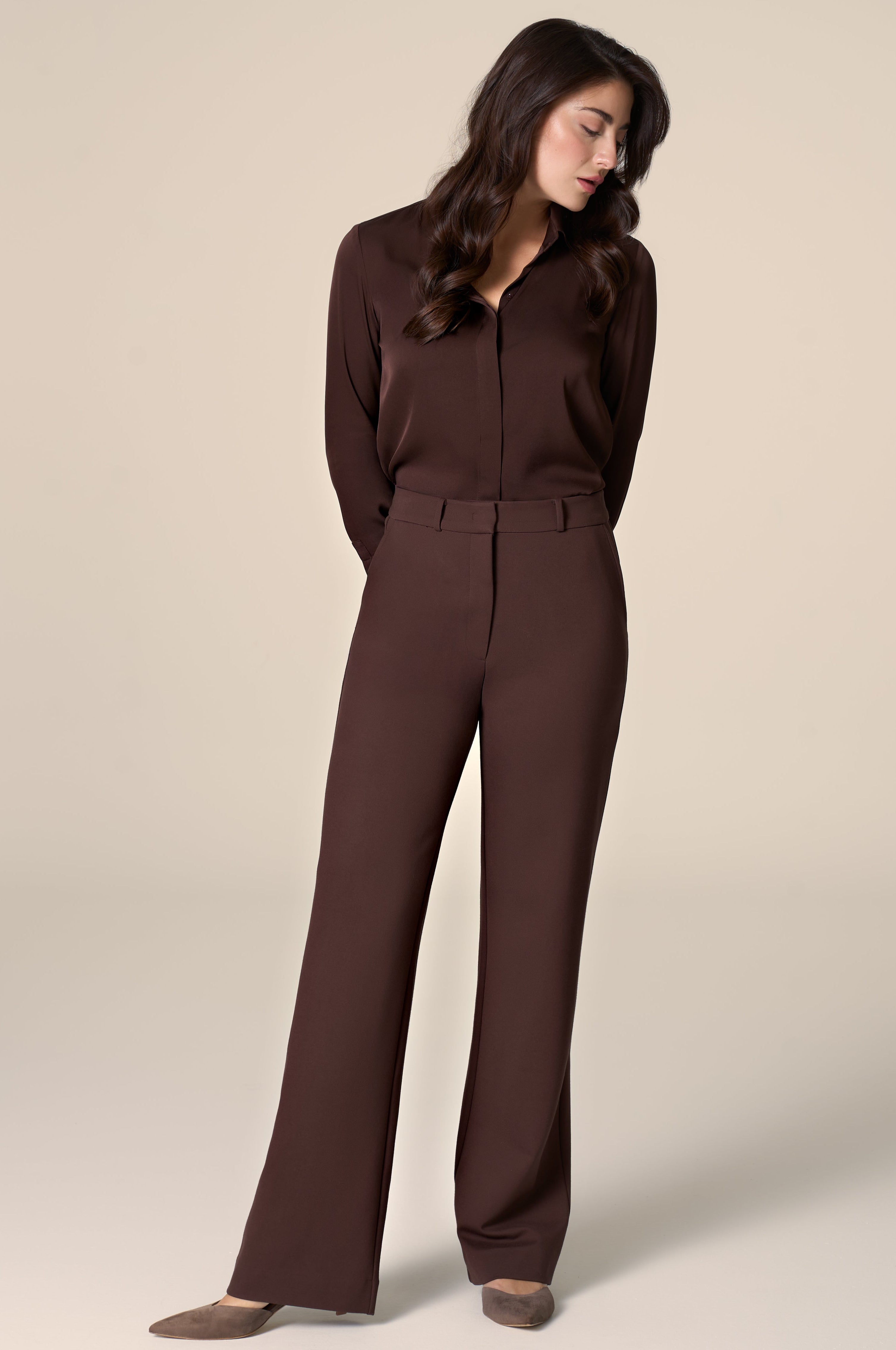 Woman wearing brown silk blouse and tailored work pants in front of a plain background.
