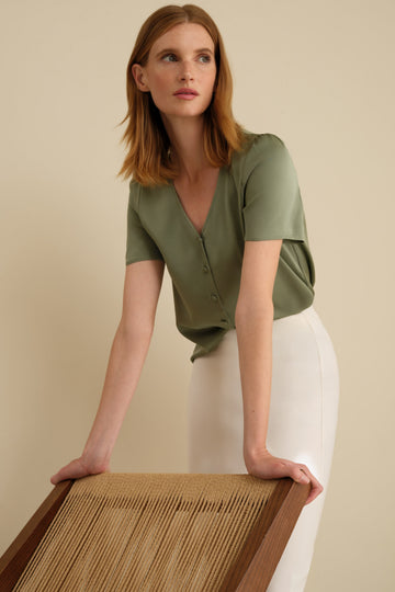 Woman standing in a white room wearing a sage green silk blouse with short sleeves and an ivory midi length pencil skirt.